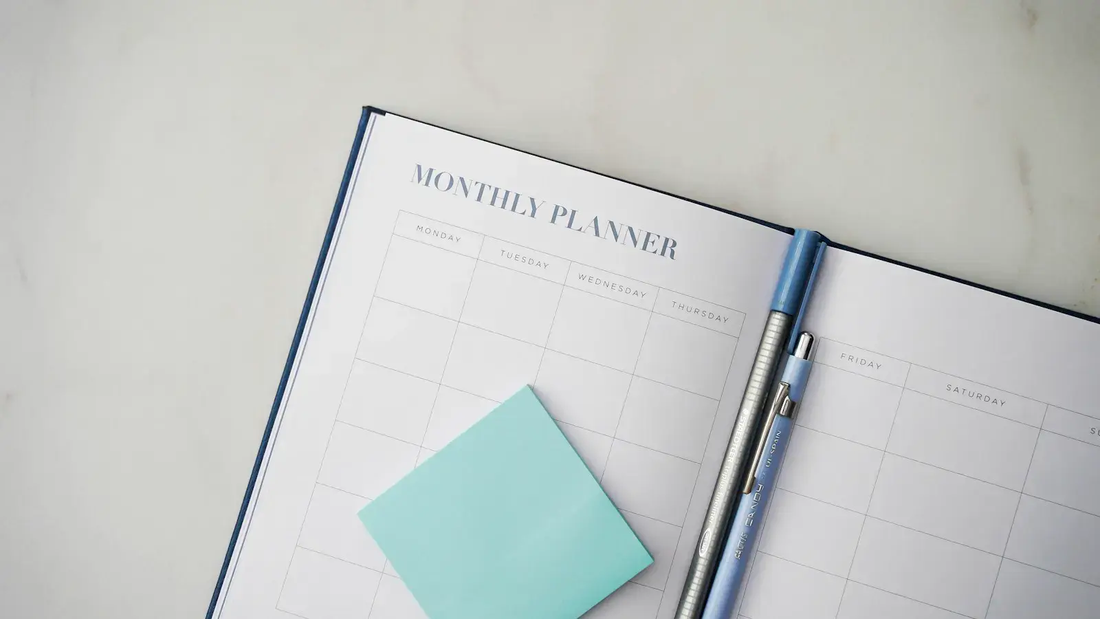 An open monthly planner with an empty grid of days, a pen, and a sticky note